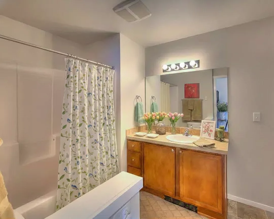 A bathroom with a floral shower curtain, double sinks with wooden cabinets, a large mirror, and a countertop decorated with flowers and framed art.